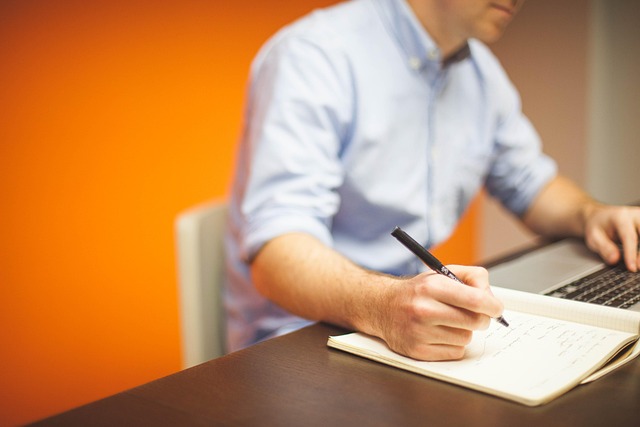 A person writing in a notebook next to a laptop